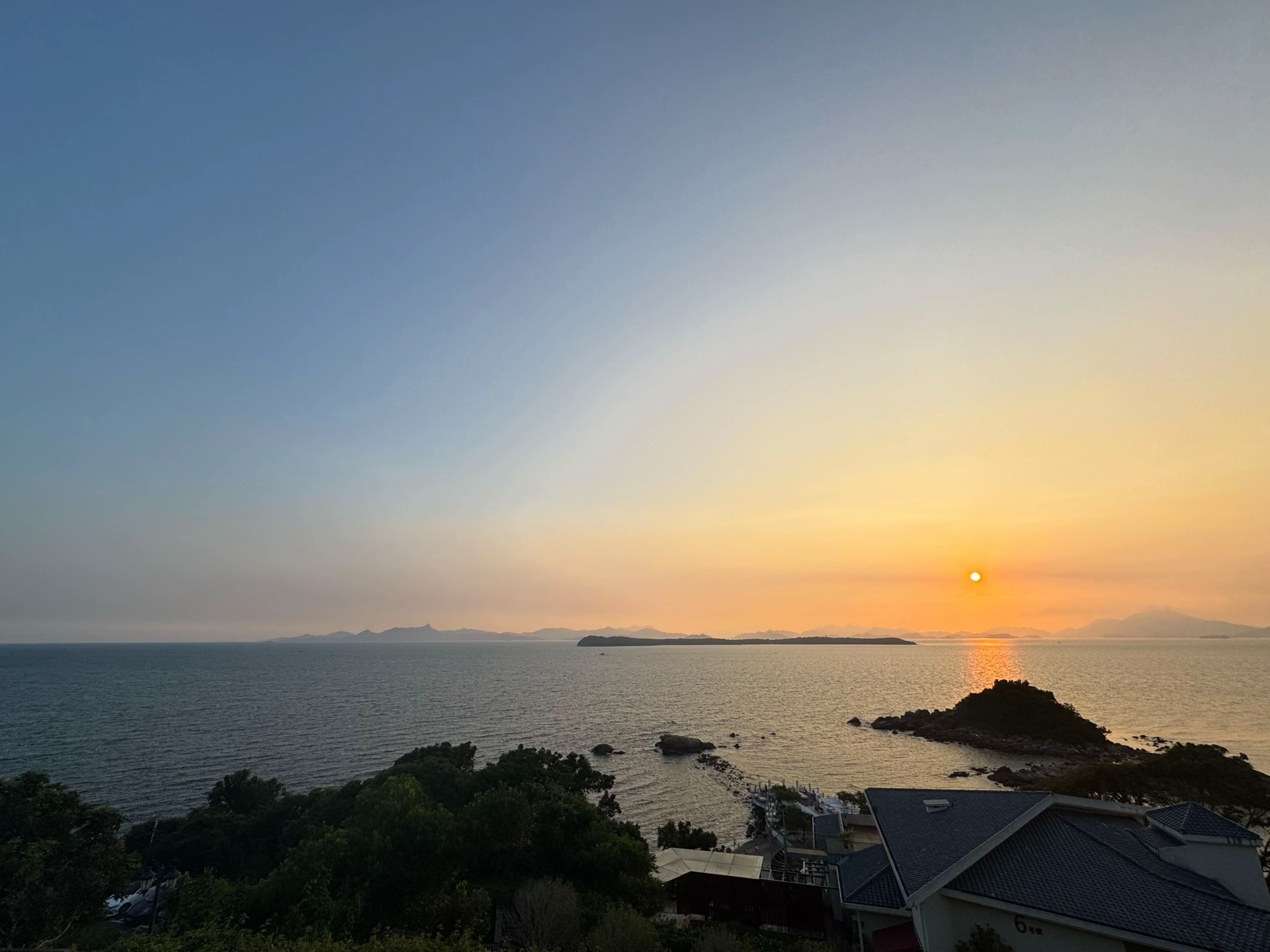 从高处俯瞰的海面，太阳正要沉入海平线，岸边露出房屋与树影