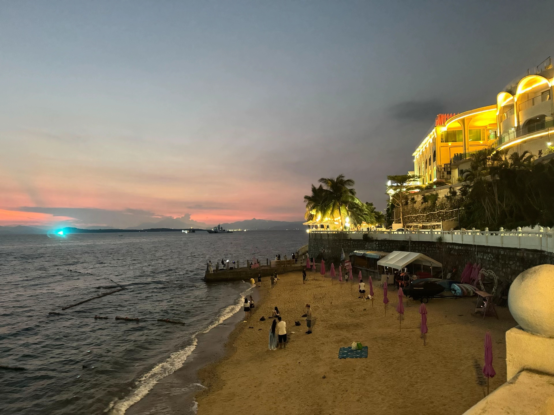 入夜后的海滩，游客在海边停留，山坡上的建筑亮着灯